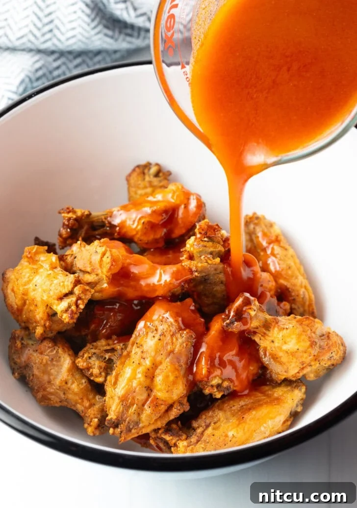 Pouring spicy buffalo sauce from a measuring cup onto fried chicken wings in a bowl.