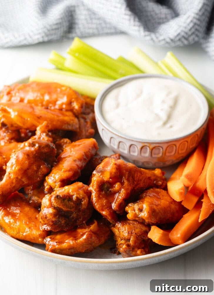 Plate of saucy buffalo chicken wings. On the plate is also a ramekin of ranch dressing, a pile of carrot sticks, and a pile of celery sticks.