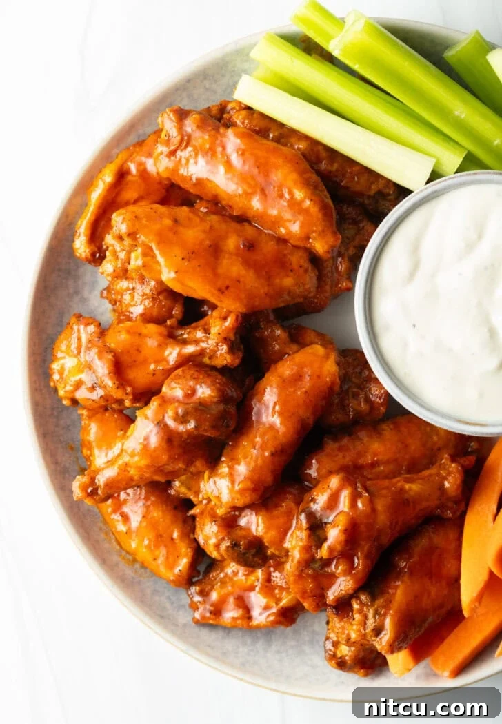 Plate of saucy buffalo chicken wings. On the plate is also a ramekin of ranch dressing, a pile of carrot sticks, and a pile of celery sticks.
