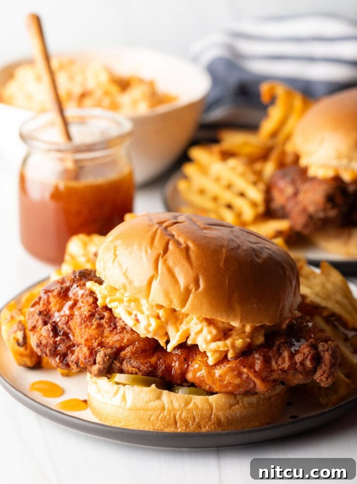 Honey pepper pimento chicken sandwich on a plate. In the background is a jar of honey, and second sandwich with waffle fries.