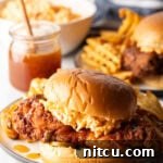 Honey pepper pimento chicken sandwich on a plate. In the background is a jar of honey, and second sandwich with waffle fries.