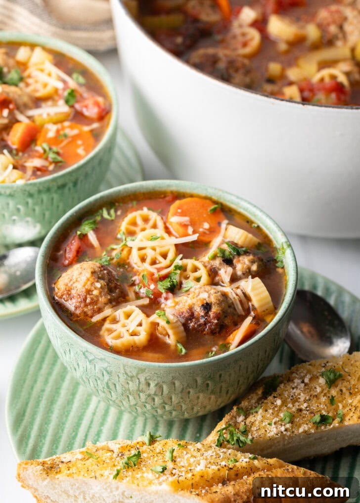 Green bowl of Italian meatball soup loaded with wheel shaped pasta, sliced carrots, in a tomato broth.