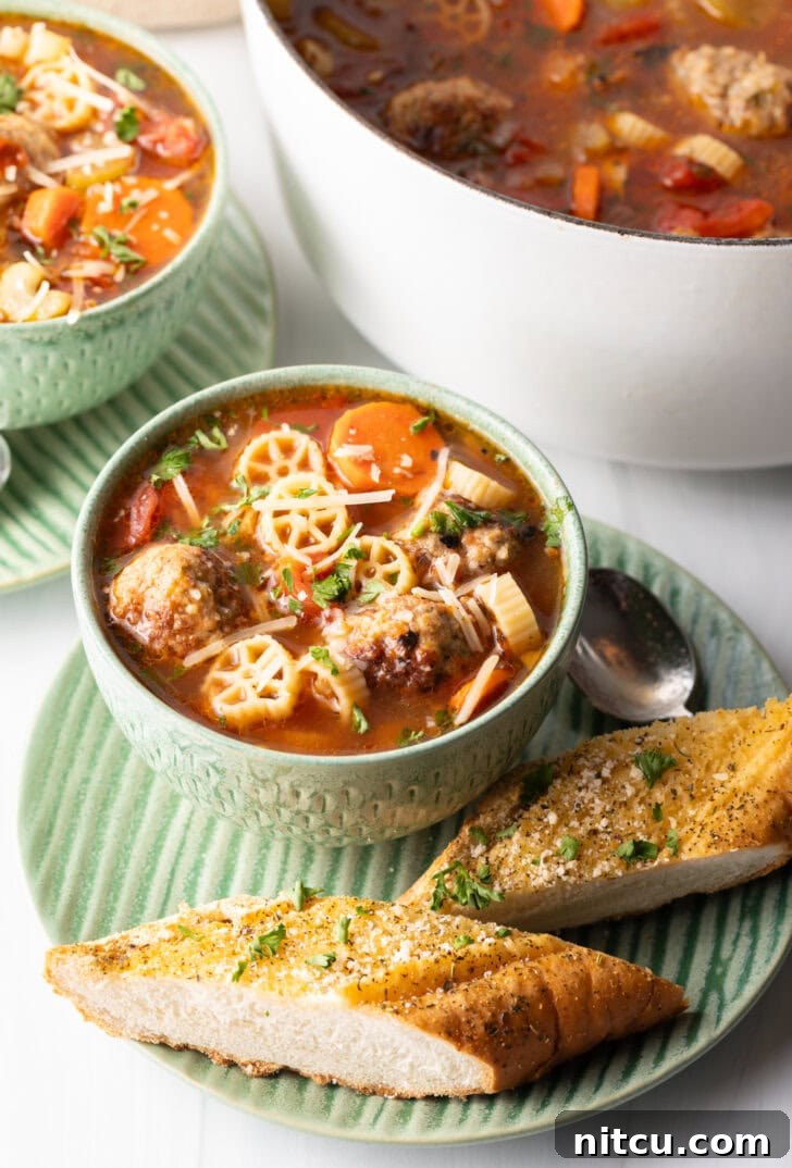 Green bowl of Italian meatball soup loaded with wheel shaped pasta, sliced carrots, in a tomato broth. Two pieces of toasted baguette are on the green plate that's beneath the soup.
