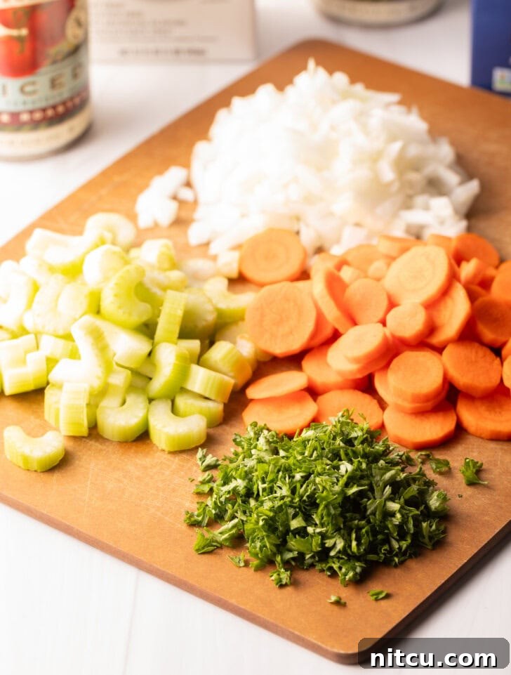 Wooden cutting board with individual piles of slices carrots, celery, diced onion, and parsley.
