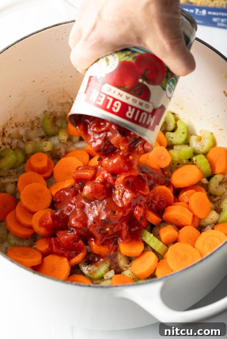 Hand adding canned tomatoes to pot with fresh slices of carrots and celery.