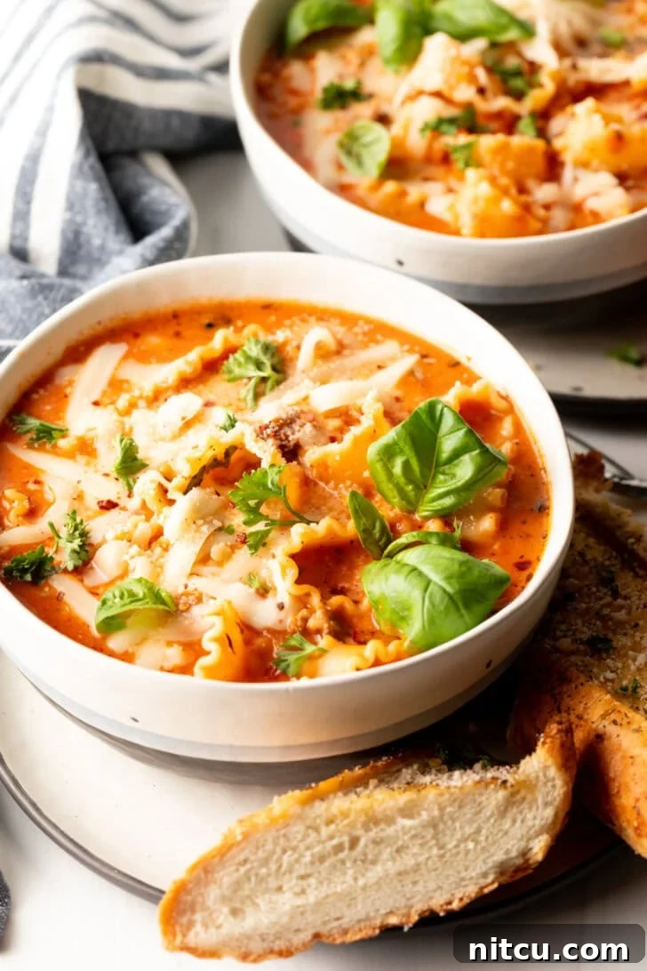 A vibrant white bowl overflowing with homemade lasagna soup, beautifully garnished with two fresh basil leaves. A rustic slice of baguette rests on a white plate beneath the bowl, ready for dipping.