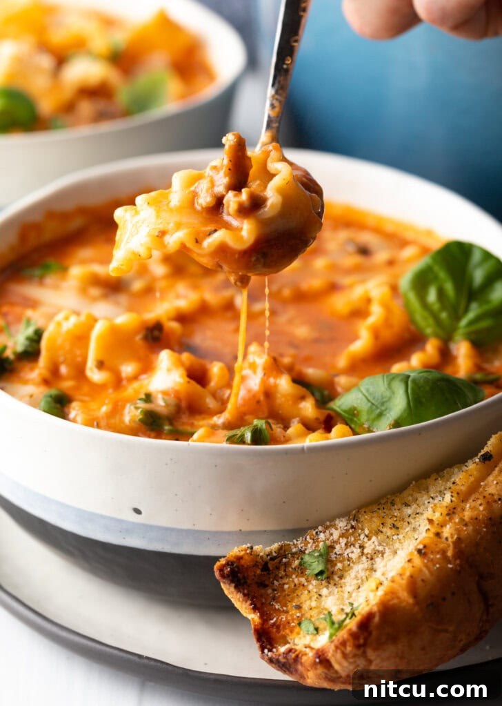 A spoon gracefully scoops a hearty bite of tomato and cheesy lasagna soup, showcasing the perfectly cooked pasta, savory sausage, and rich broth.