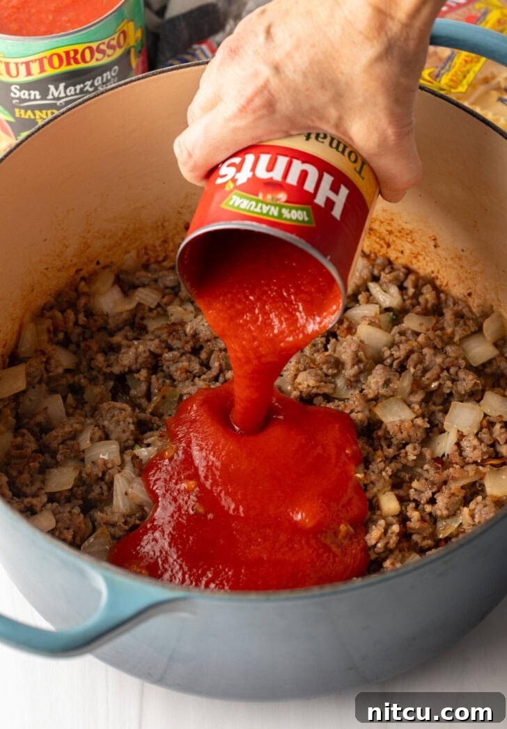 A hand gently adds canned tomato sauce to a pot of cooked sausage and onions, enriching the soup's tomato base.