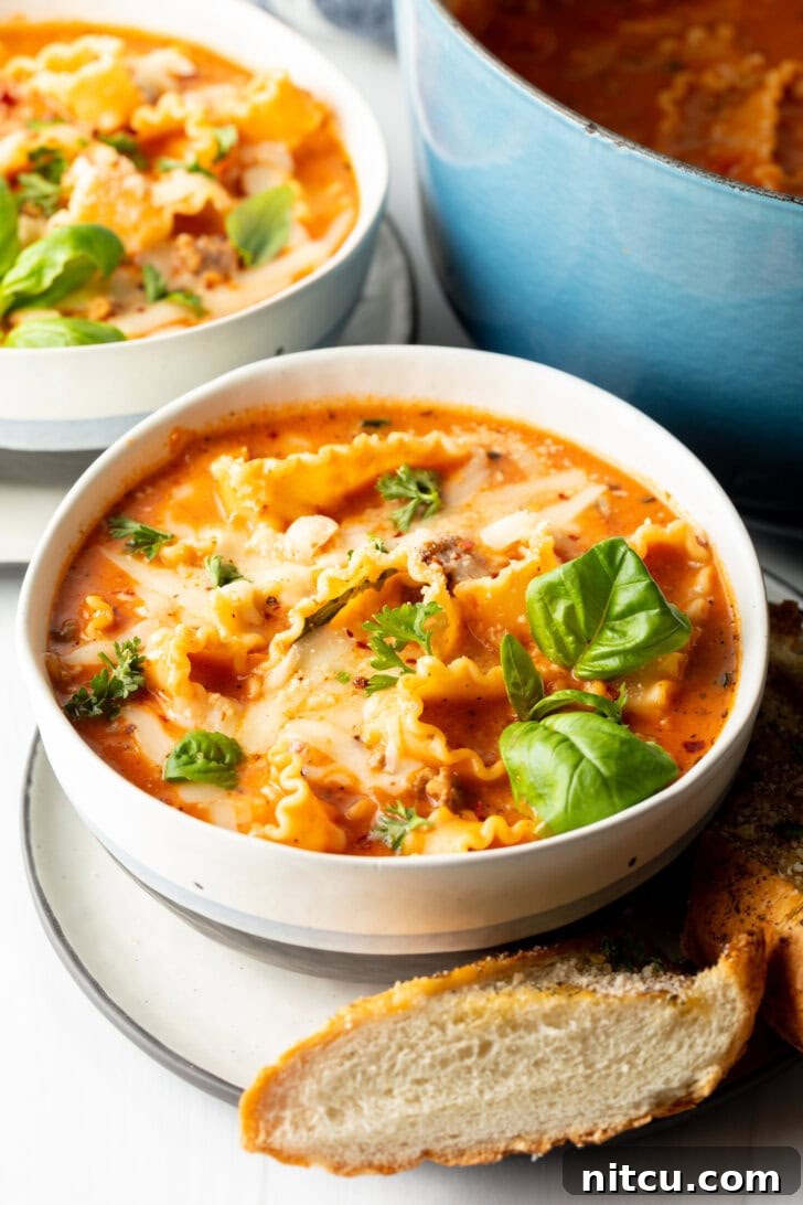 A inviting white bowl of lasagna soup, garnished with vibrant fresh basil leaves, with a side of toasted baguette on a white plate, ready to be enjoyed.