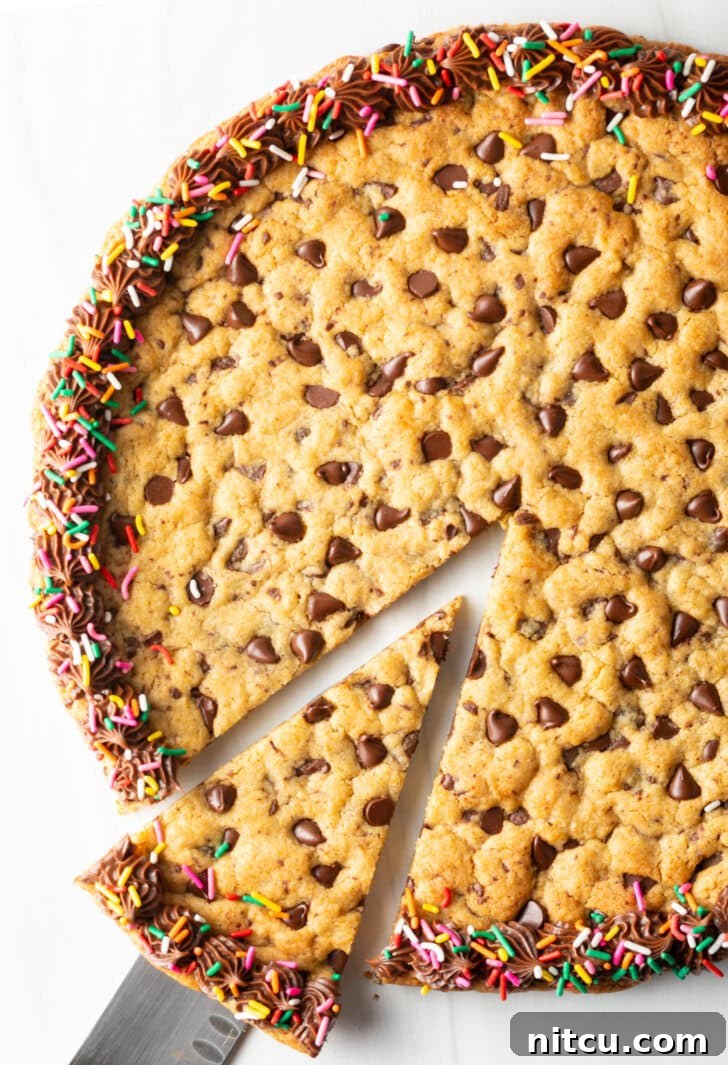 Large chocolate chip cookie cake. The outside edge is piped with chocolate frosting and covered with rainbow sprinkles. A cake cutter is taking a wedge slice from the cake.