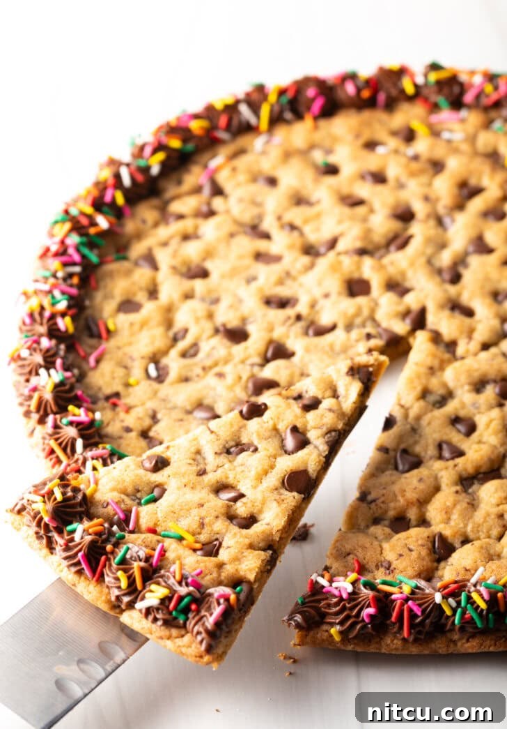 Large chocolate chip cookie cake. The outside edge is piped with chocolate frosting and covered with rainbow sprinkles. A cake cutter is taking a wedge slice from the cake.