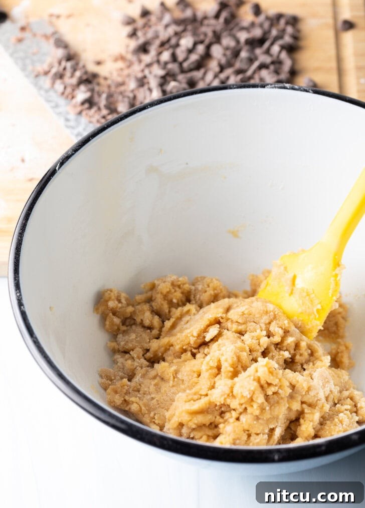 White mixing bowl with cookie dough ingredients, stirred by a yellow spatula.