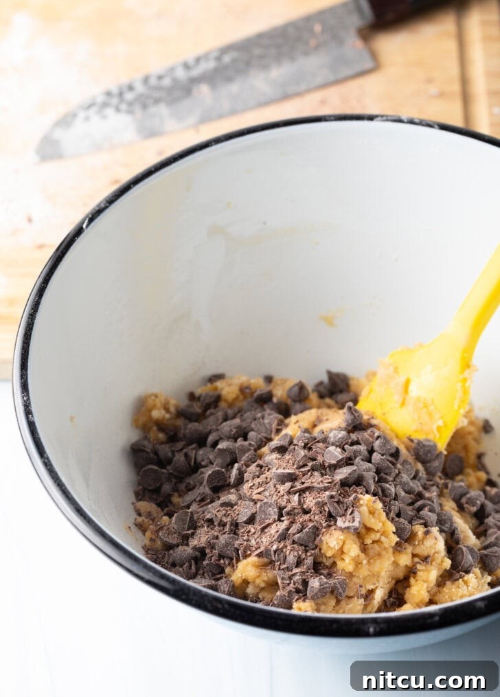 White mixing bowl with cookie dough ingredients, stirred by a yellow spatula.