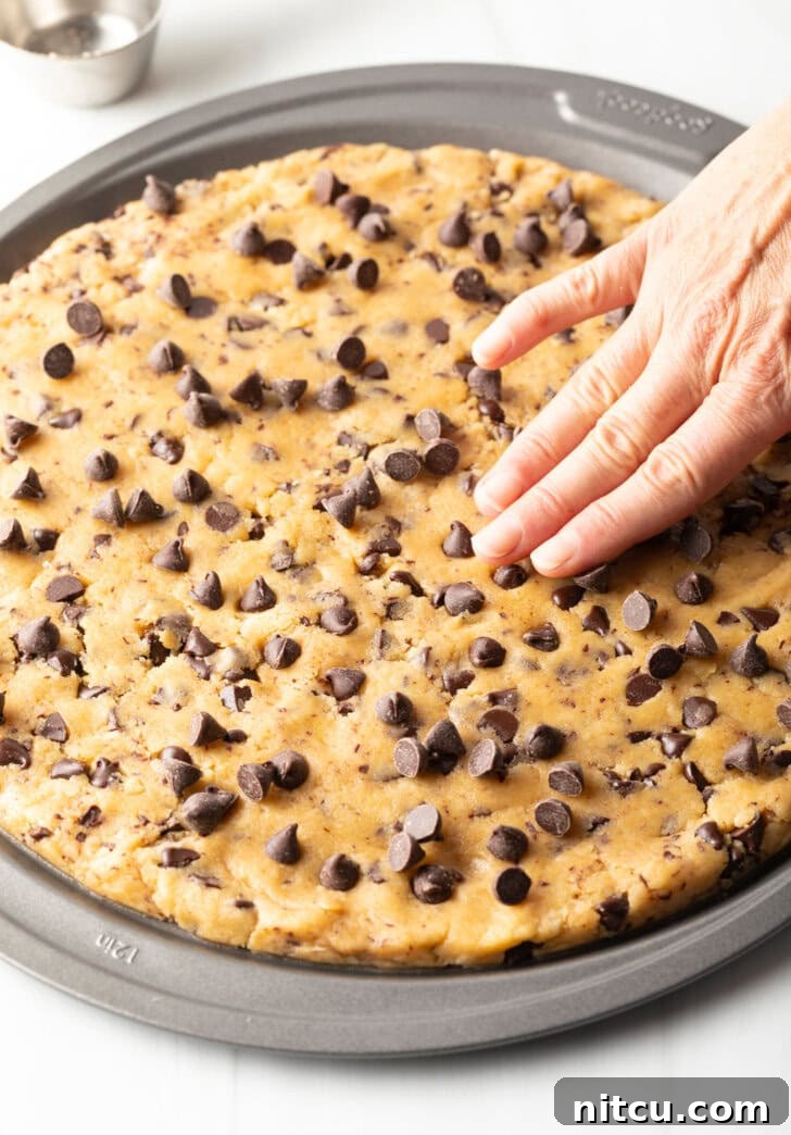 Large chocolate chip cookie cake, hand spreading dough out in a pizza pan.