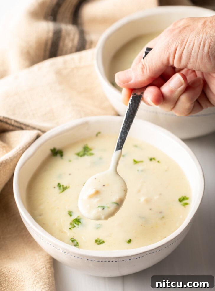 Top-down view of a white bowl with creamy chicken soup garnished with fresh parsley. A hand holds a metal spoon with a spoonful of soup for the camera, emphasizing its rich texture.