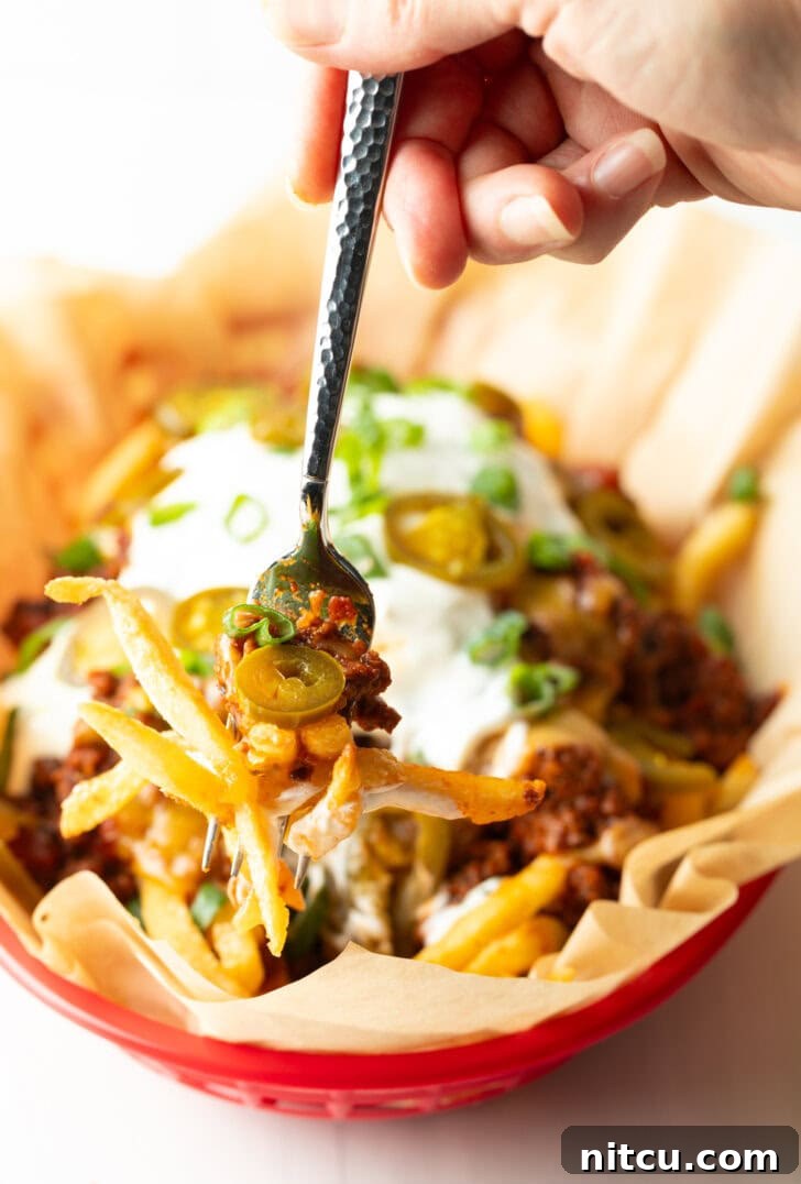 A rustic basket lined with parchment paper, overflowing with golden, crispy french fries. They are generously smothered with a rich, hearty chili, and topped with a glistening layer of melted cheddar cheese. A dollop of cool sour cream, a scattering of fresh chopped green onions, and vibrant sliced jalapeños complete the presentation. A hand holding a forkful of this delicious creation is visible, ready to enjoy.
