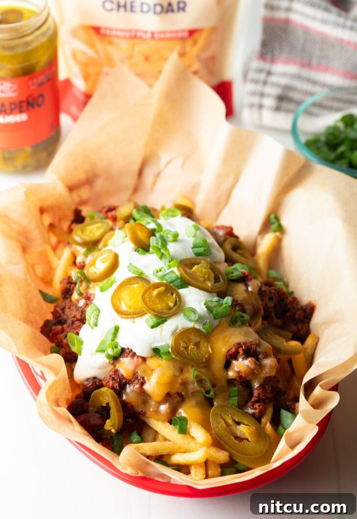 A rustic basket lined with parchment paper, bursting with crispy french fries lavishly topped with rich chili and melted cheddar cheese. It's elegantly garnished with creamy sour cream, vibrant chopped green onions, and fiery sliced jalapeños, showcasing a truly indulgent dish.