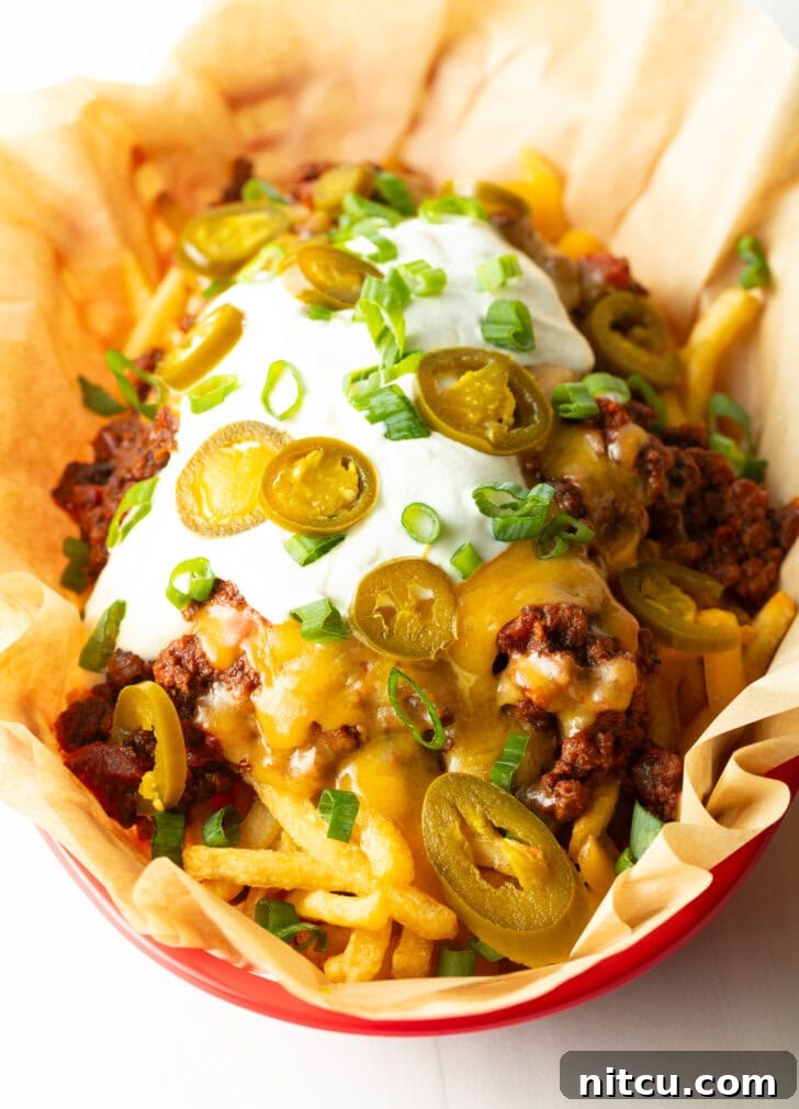 A rustic basket lined with parchment paper, filled with crispy french fries generously loaded with rich chili, melted cheddar cheese, cool sour cream, fresh chopped green onions, and vibrant sliced jalapeños. The dish is presented beautifully, ready to be enjoyed.