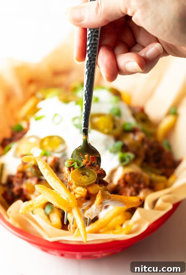 A rustic basket lined with parchment paper, filled with crispy french fries lavishly topped with rich chili, perfectly melted cheddar cheese, cool sour cream, fresh chopped green onions, and vibrant sliced jalapeños. A hand holding a forkful, showcasing the delicious layers, is visible in the foreground.