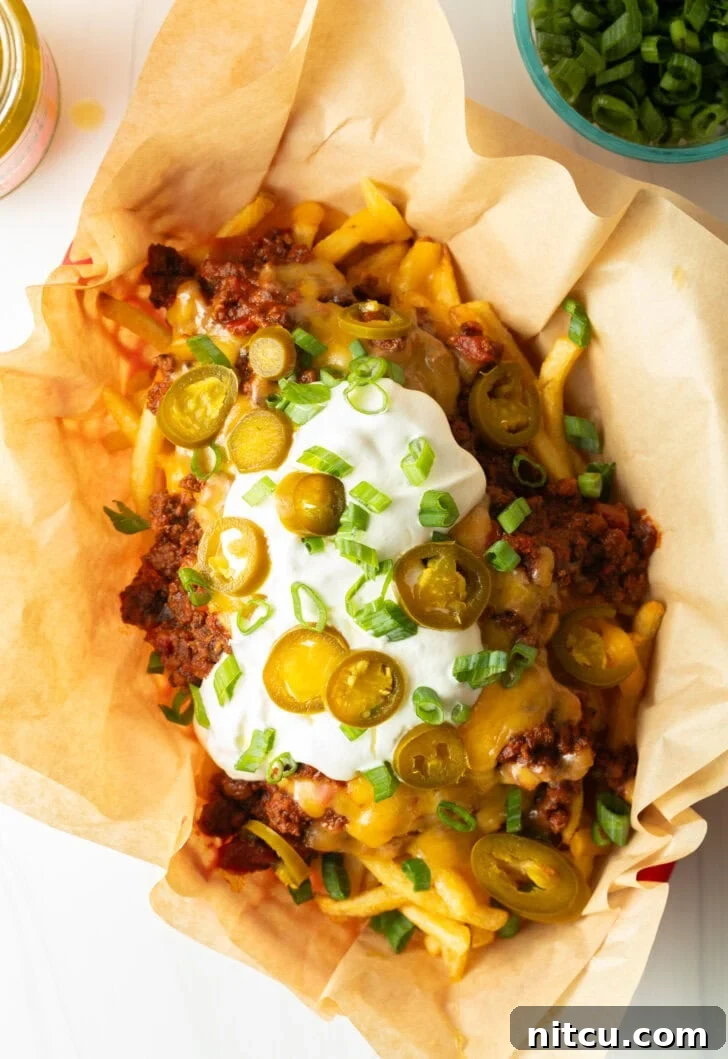A rustic basket lined with parchment paper, filled with crispy french fries generously covered with rich chili and a thick layer of melted cheddar cheese. It's garnished with cool sour cream, vibrant chopped green onions, and fiery sliced jalapeños, ready to be served.