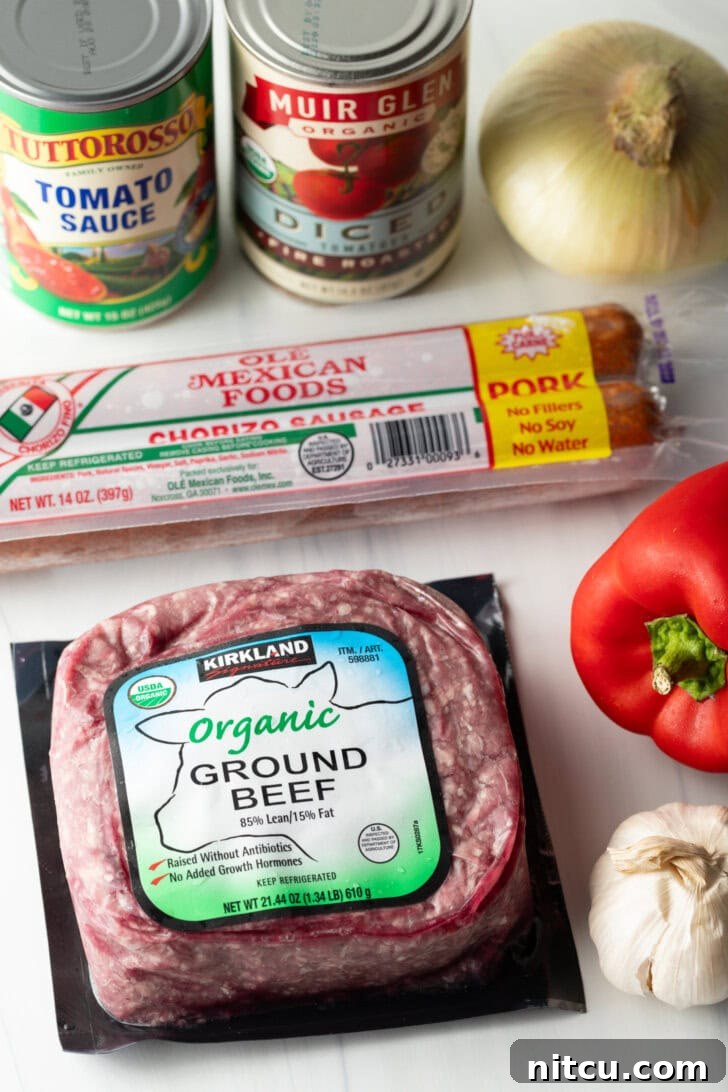 A curated display of raw ingredients essential for making the homemade chili. This includes a package of fresh ground beef, a portion of vibrant Mexican chorizo, a can of rich tomato sauce, a can of plump diced tomatoes, a whole crimson red bell pepper, and a complete bulb of fragrant garlic.
