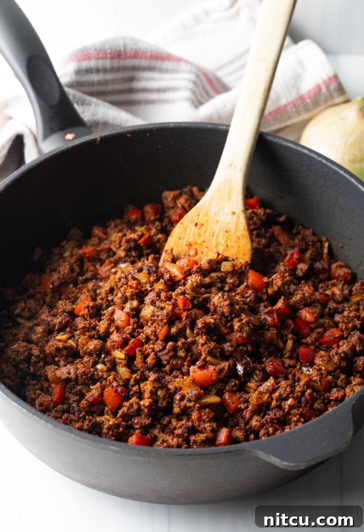 A wooden spoon stirring a vibrant, simmering chili in a deep, black cast iron skillet. The chili is rich with meat, vegetables, and spices, indicating it's in the process of developing deep flavors.