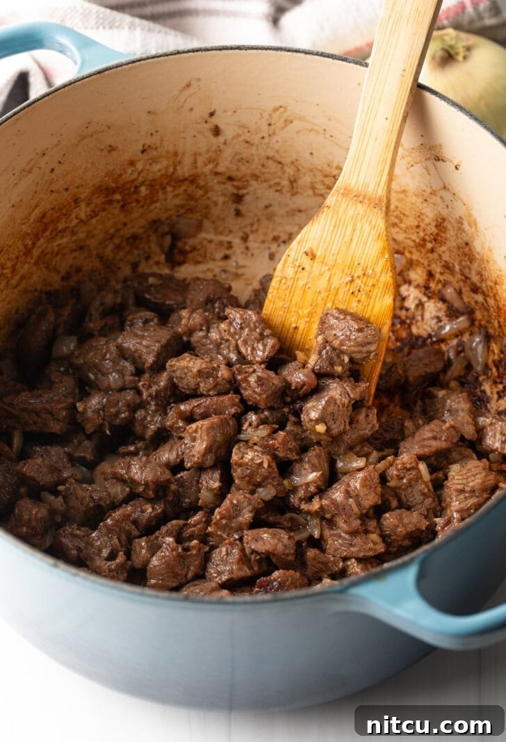 Wood spatula stirring pieces of beef as they cook in a large pot, alongside chopped onions.