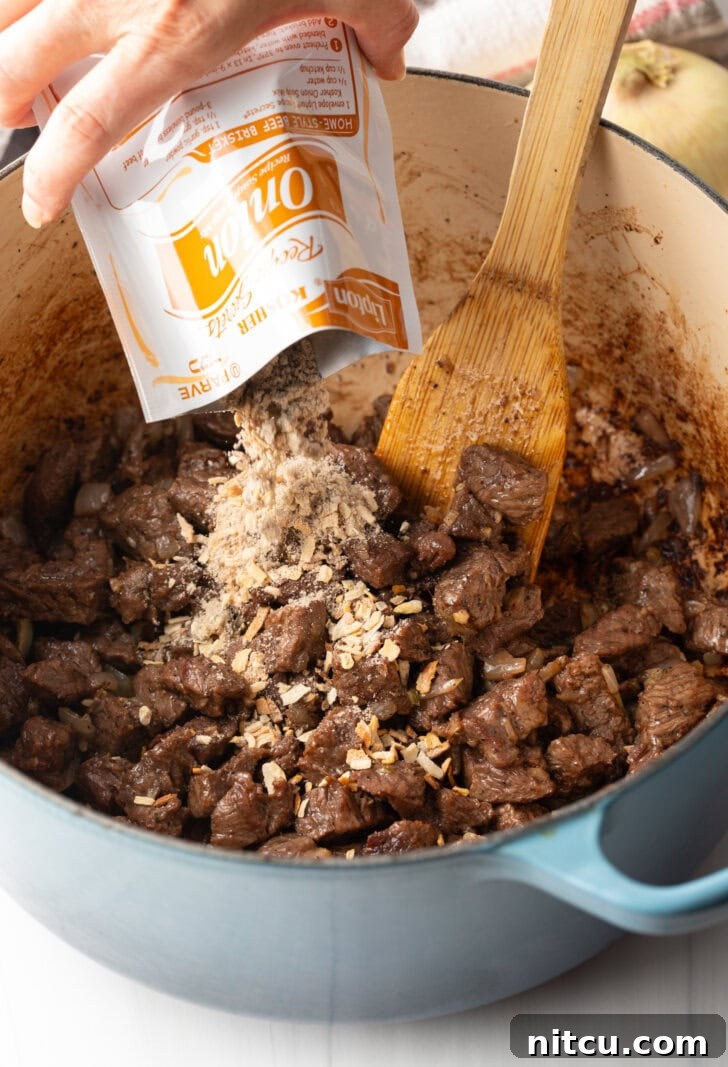 Adding seasonings from an onion soup packet to the cooked beef and onions in a large pot, with a wood spatula.