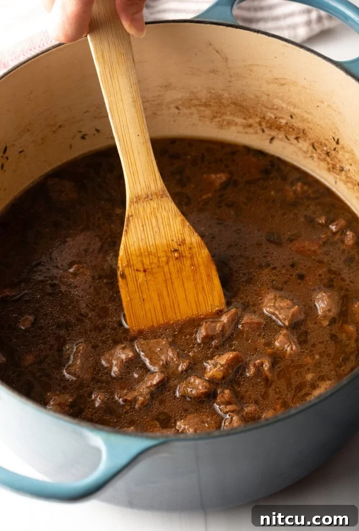 Wooden spoon stirring pieces of cooked beef in a rich gravy in a large cooking pot.