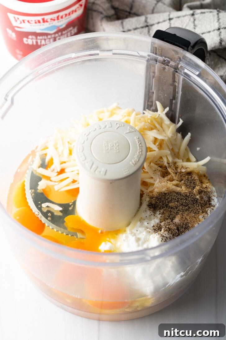 Various ingredients including shredded cheese, cottage cheese, whole eggs, and spices are placed inside a food processor, ready for blending.