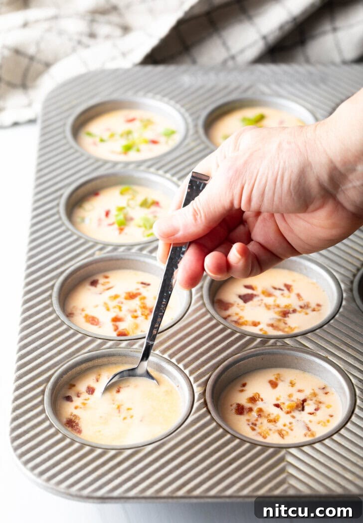 A hand uses a metal spoon to add the blended egg mixture to individual cups in a metal muffin pan.