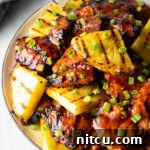 Top down view of sticky chicken pieces topped with chopped scallions and grilled slices of pineapple.