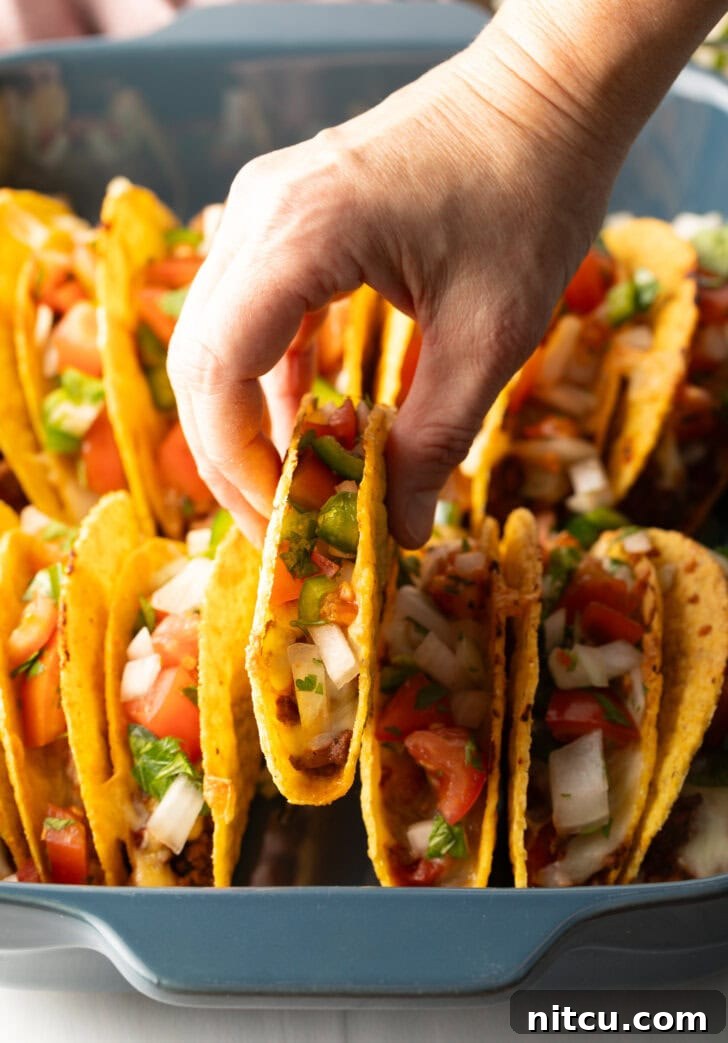 A hand picking up a single taco from the baked array in the tray, showcasing its crispy shell and generous filling.