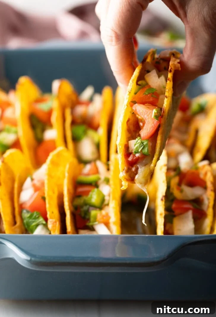 A hand reaching into a baking dish to pick up one of the freshly baked, loaded tacos.
