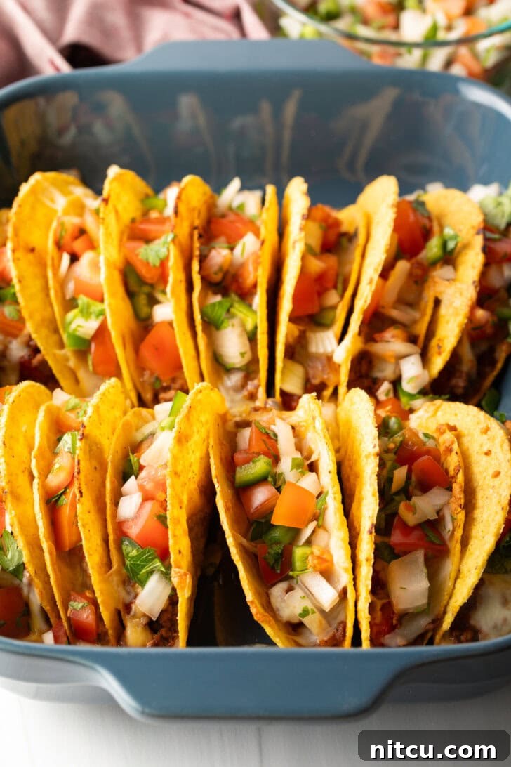Two rows of a dozen loaded yellow corn hard shell tacos, standing upright in a large baking dish, ready for the oven.