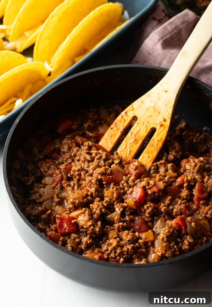 Wooden spatula stirring ground beef taco mixture in a large black deep skillet, browning evenly.