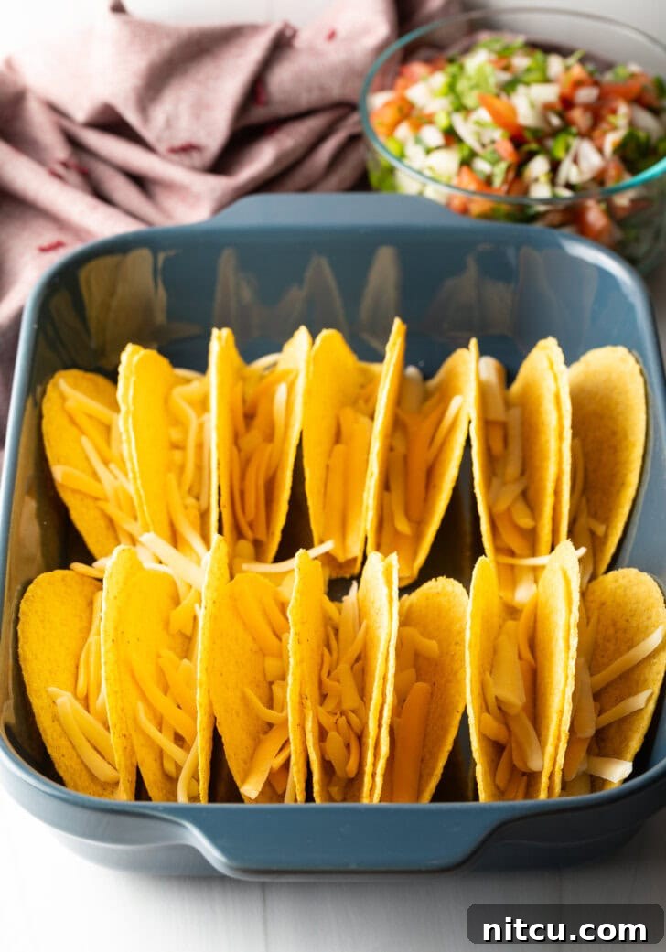 Two rows of a dozen yellow corn hard shell tacos, standing upright in a large baking dish, with shredded cheese sprinkled in the bottoms.