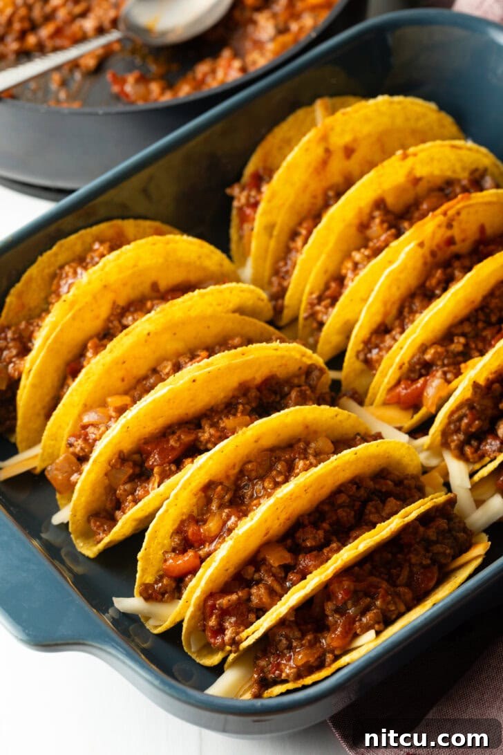 Ground beef mixture being added to yellow corn tortilla shells arranged in a baking dish.