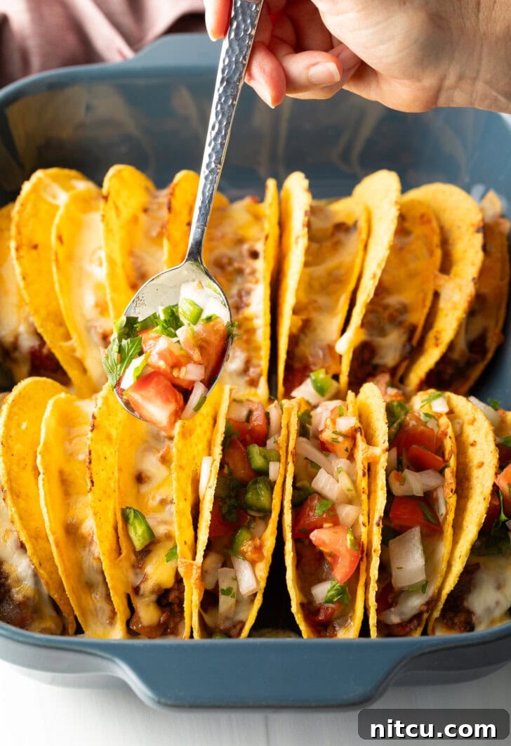 A spoon adding fresh pico de gallo to a row of hard shell tacos, nestled in a baking dish.