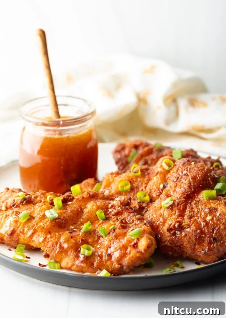 Three pieces of golden-brown, fried hot honey chicken on a plate, garnished with vibrant sliced green onions. A jar of golden honey with a wooden dipper is visible in the background.