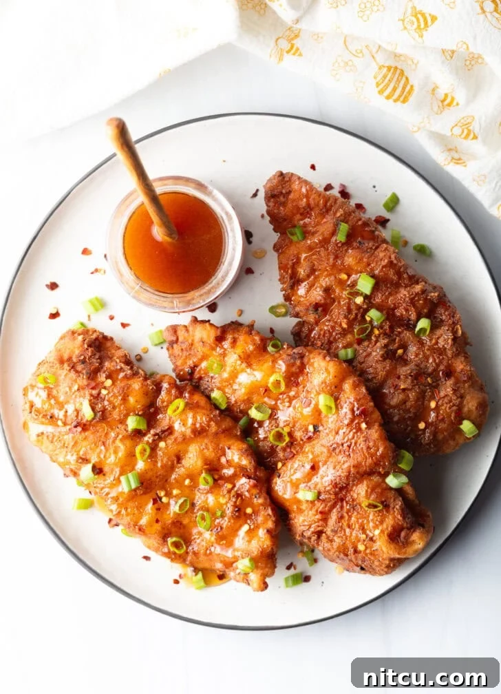 A delicious top-down view of three hot honey fried chicken pieces, generously garnished with green onions. A rustic wooden spoon rests in a jar of honey in the background.