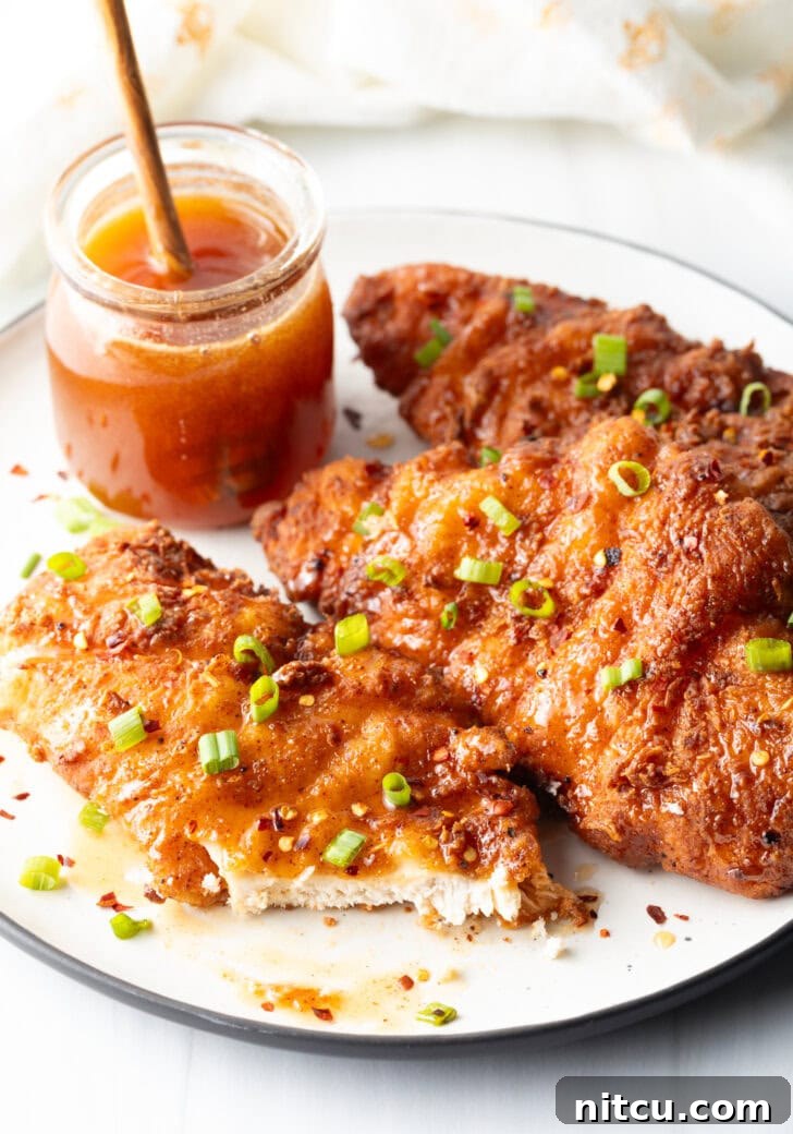 Three pieces of fried hot honey chicken, exquisitely plated and garnished with sliced green onions. A jar of honey sits in the background, and the front piece of chicken is cut open, revealing its tender, juicy interior.
