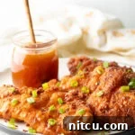 A close-up of three pieces of fried hot honey chicken, beautifully arranged on a plate with fresh green onions. A jar of honey is present in the background.
