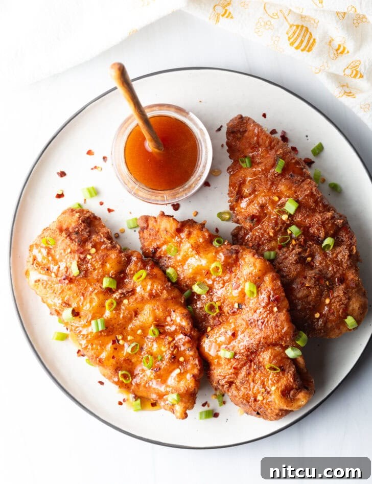 A top-down perspective of three glistening pieces of fried hot honey chicken, adorned with fresh green onions. A jar of honey and a wooden spoon are softly blurred in the background.
