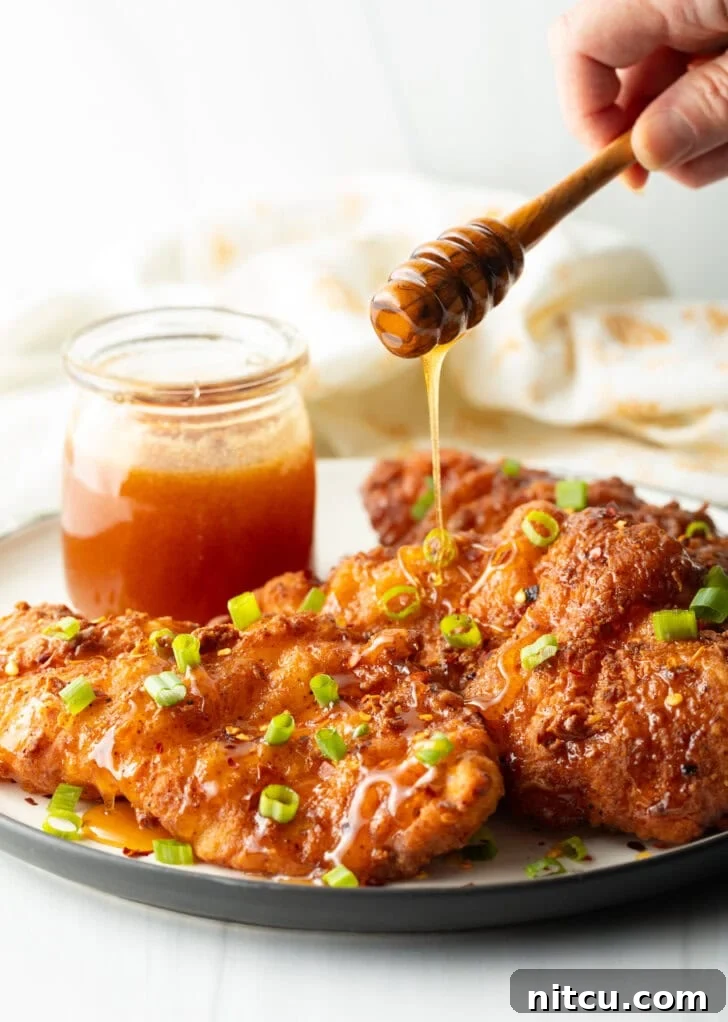 A hand expertly drizzling a generous stream of golden hot honey sauce over three pieces of freshly fried chicken, with green onions and a honey jar in the frame.