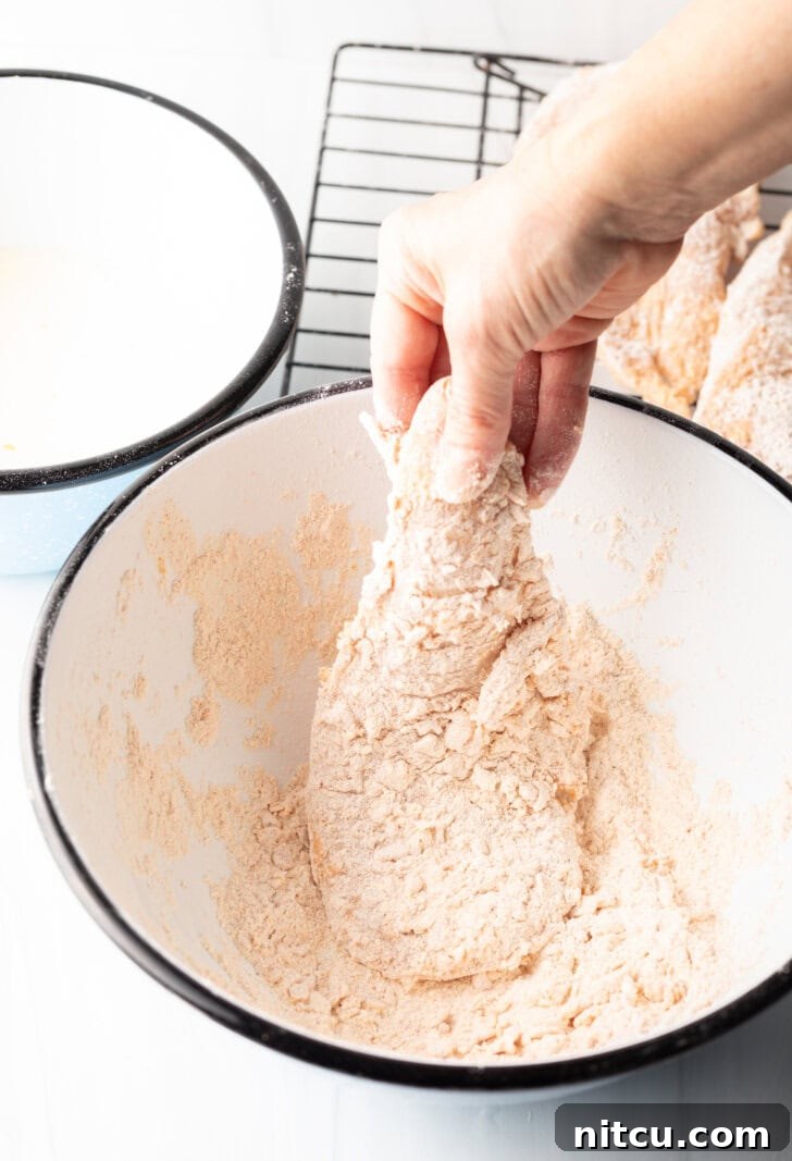 A hand gently coating a raw chicken breast in a bowl of white flour, demonstrating the initial dredging step for even coverage.