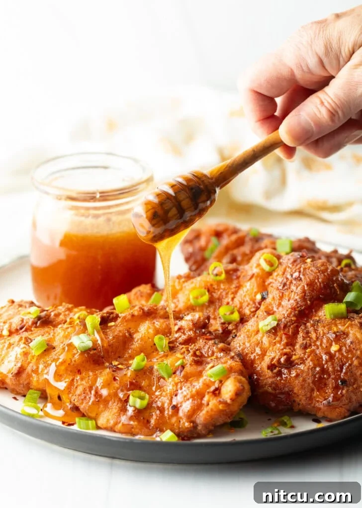 A mesmerizing shot of hot honey sauce being drizzled over three perfectly fried chicken pieces, garnished with green onions. A jar of honey is prominently featured in the background.
