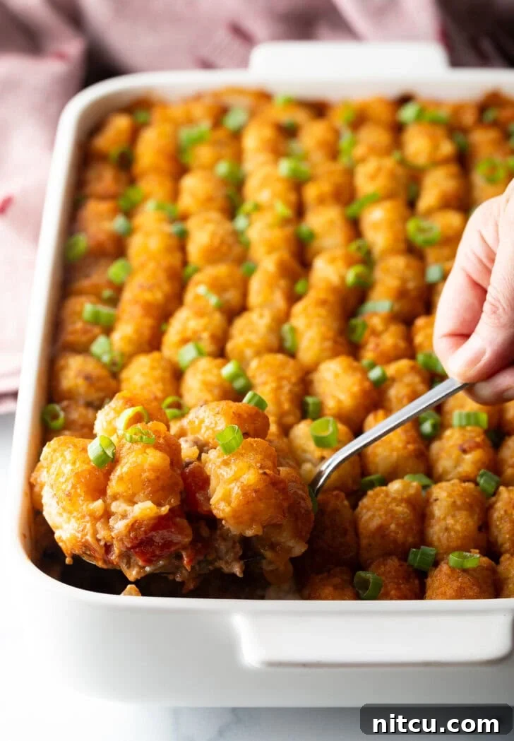 A fully baked Cowboy Tater Tot Casserole, showcasing rows of perfectly golden tater tots on top. A hand with a metal spoon scoops a generous serving from the dish.
