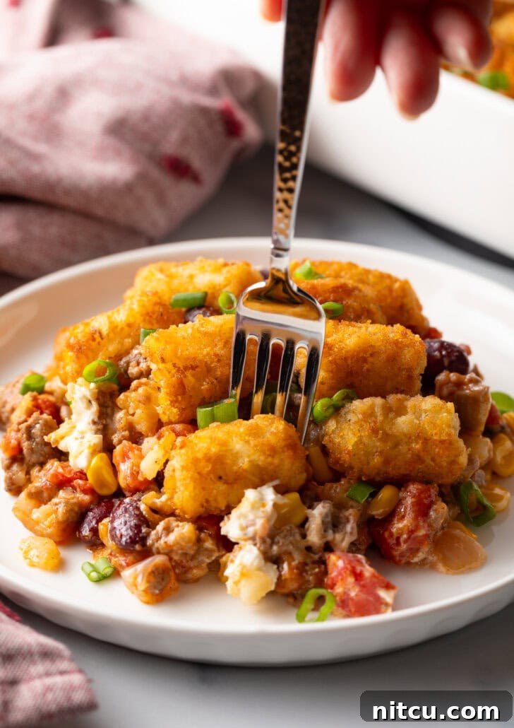 White plate with tater tot and ground beef cowboy casserole. A fork is taking a bite from the plate, showing the creamy, meaty filling.