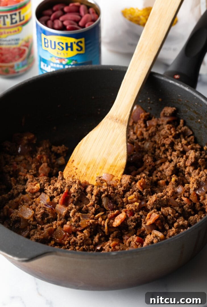 Deep black skillet with ground beef mixture, being stirred by a wooden spatula.